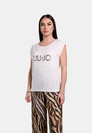 Donna con capelli lunghi e scuri che indossa un top bianco senza maniche con la scritta "LIU·JO" e pantaloni a gamba larga con stampa animalier, in piedi davanti a uno sfondo semplice.