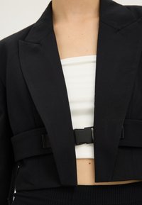 Tät bild av en person som bär en svart kort blazer med breda slag och en framfästning över en vit kort topp.