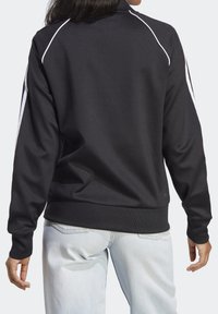 Person som bär en svart långärmad jacka med vita ränder på ärmarna och ljusblå jeans, visad bakifrån.