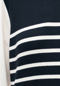 Tissu tricoté bleu marine avec des rayures blanches horizontales, à côté d'un panneau de tissu blanc lisse, montrant un gros plan d'un vêtement.