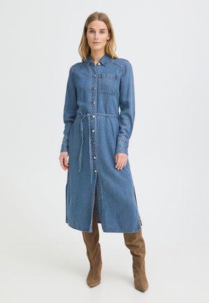 Femme portant une robe en jean à manches longues boutonnée avec une poche poitrine et une ceinture, associée à des bottes marron en daim montant jusqu'aux genoux, debout sur fond blanc.