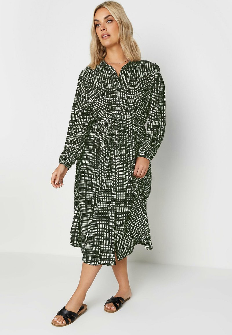 Grön mönstrad klänning med långa ärmar, knäppning framtill, elastisk midja och en flödande kjol; i kombination med svarta sandaler.