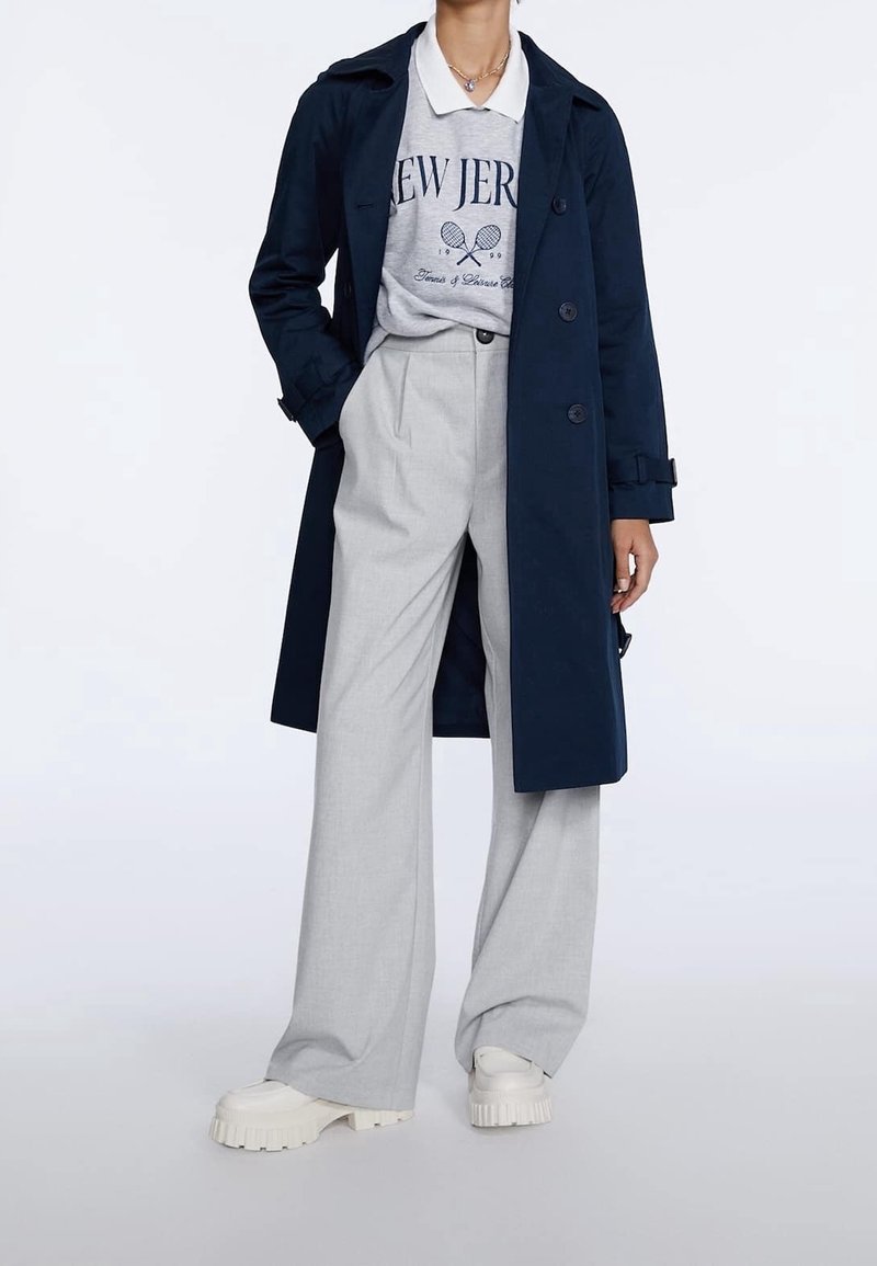 Un manteau trench bleu marine sur un sweat-shirt gris avec l'impression 'New Jersey', associé à un pantalon large gris et des chaussures à plateforme blanches et épaisses.