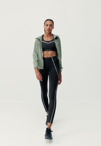 Športový outfit pozostávajúci z svetlozeleného bundy, čierneho športového podprsenka a čiernych legín s bielymi bočnými pruhmi. Čierne športové topánky dokončujú celý vzhľad.