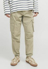 Beige Cargo-Hosen mit elastischen Bündchen, Seitentaschen und einem lockeren Schnitt. Kombiniert mit braunen und cremefarbenen Sneakers sowie einem gestreiften Langarmshirt.