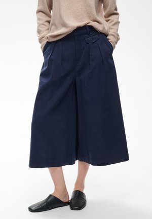 Personne portant un pantalon large cropped bleu marine, un pull beige et des chaussures plates noires slip-on sur un fond clair uni.