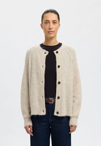 Beige wollen cardigan met een ronde halslijn, vijf donkere knopen, ribbelstructuur en verlaagde schouders. Gestyled over een donkere top en jeans.