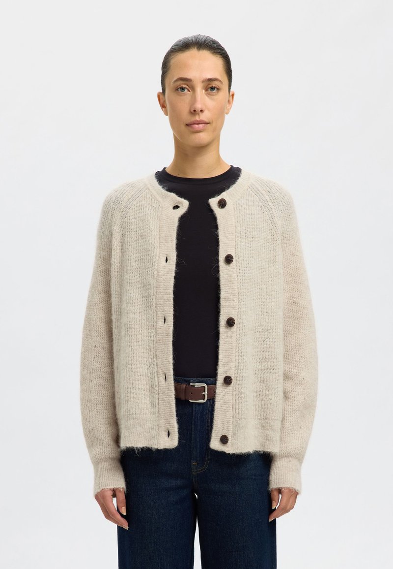 Beige wollen cardigan met een ronde halslijn, vijf donkere knopen, ribbelstructuur en verlaagde schouders. Gestyled over een donkere top en jeans.