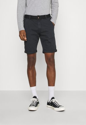 Homme portant un short noir retroussé avec ceinture noire, chemise grise à manches longues, chaussettes blanches et baskets basses noires, debout.