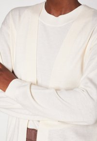 Cardigan crème clair avec ouverture devant, manches longues et poignets côtelés. Texture douce, associé à un haut beige.