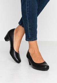 Schwarze Lederpumps mit einer runden Zehenpartie, niedrigem Blockabsatz und glatter Textur, kombiniert mit dunklen Jeans, in einer Nahaufnahme.