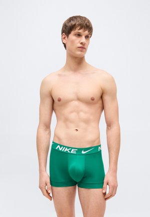 Noor mees, kellel on lühikesed pruunid juuksed, seisab ilma särgita ja kannab rohelist Nike bokserpükste, taust on hele ja lihtne.