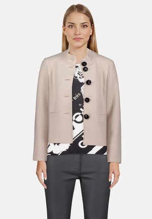 Jasno beżowa strukturalna kurtka z okrągłym kołnierzem, pięcioma czarnymi guzikami i bocznymi kieszeniami, noszona na czarno-białej wzorzystej bluzce.