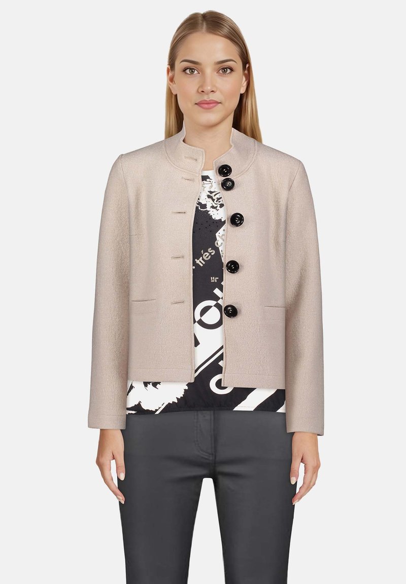 Jasno beżowa strukturalna kurtka z okrągłym kołnierzem, pięcioma czarnymi guzikami i bocznymi kieszeniami, noszona na czarno-białej wzorzystej bluzce.