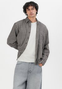 Bruine en grijze geruite button-up shirt met een relaxte pasvorm over een witte crew neck t-shirt, gecombineerd met lichtgrijze jeans.