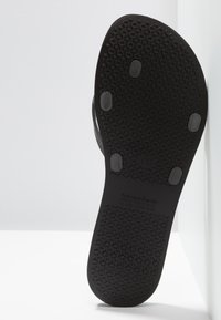 Schwarze Gummisandalen-Sohle mit einem strukturierten Grip-Muster, das glatte Kanten und erhabene Punkte für besseren Halt aufweist. Markenname auf der Oberfläche eingeprägt.