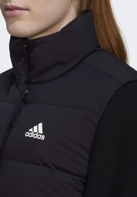 Zwart gewatteerd vest met hoge kraag, drukknoopsluiting en een wit Adidas-logo op de linkerborst. Gladde textuur en gestructureerd ontwerp.