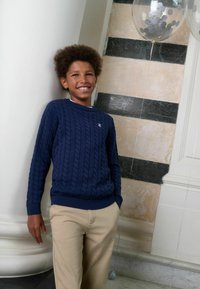 Tumesinine soonikkoes kampsun soonikute jahemate käiste ja alumise servaga, mida kannavad beežid püksid. Omaduseks on tekstuuriga mustriga ning väikese logo rinnal.