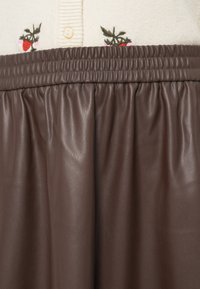 Jupe en simili cuir marron avec une taille froncée, ornée de fraises et feuilles brodées, portée au-dessus d'un cardigan crème.