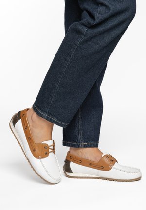Witte leren loafers met beige accenten en veters, gedragen met donkerblauwe jeans door een persoon staand op een witte achtergrond.