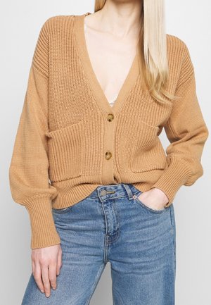 Un modèle porte un cardigan beige en grosse maille, avec des boutons et des poches plaquées, sur un jean bleu. Une main est dans une poche.