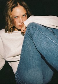 Jeune femme portant un sweat léger et un jean bleu, assise les genoux relevés, reposant son menton sur sa main, regardant droit devant elle.