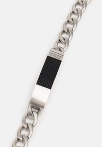 Bracelet en chaîne en acier inoxydable argenté avec un maillon central rectangulaire en caoutchouc noir présentant un motif texturé et un logo gravé.
