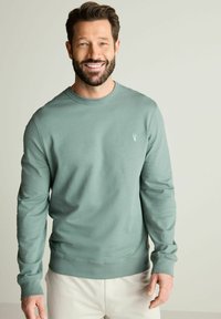Sudadera verde claro de tela suave, con cuello redondo, mangas largas y un pequeño logo bordado en el pecho.