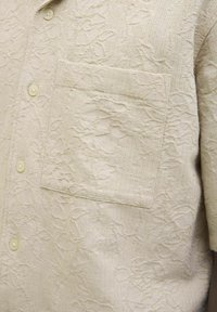 Camicia corta beige chiaro con maniche corte, caratterizzata da un motivo floreale testurizzato, chiusura con bottoni e taschino sul lato sinistro del petto.