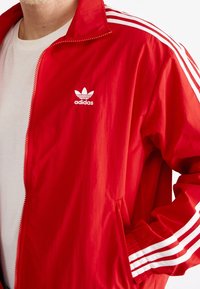 Röd zip-up jacka i lättviktsmaterial, med vitt Adidas-logotyp och tre vita ränder längs ärmarna. Elastiska muddar och midja.