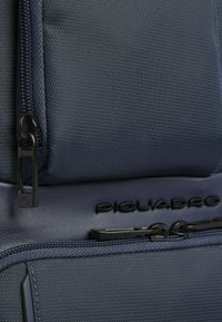 Donkerblauwe rugzak met gestructureerde stof, voorzien van een ritssluitingvak en het opgelegde "Piquadro"-logo. Zwarte hardware accenten zijn zichtbaar.