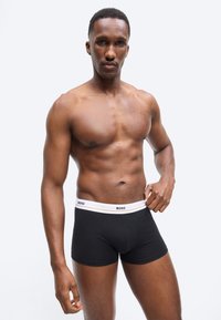 Izmos férfi, amint a "BOSS" feliratú, fehér derékpántos fekete boxermadarékját igazítja, egyszínű világos háttér előtt állva.