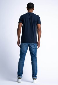 Námorně modré bavlněné tričko, krátké rukávy, kulatý výstřih. Nosené s světle modrými džínami, s ohrnutými manžetami, a bílými teniskami. Pohled zezadu.