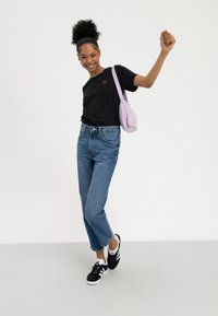 Mladi žena koja se smiješi, nosi crnu majicu s vezenim trešnjama, plave traperice, crno-bijele tenisice i nosi ljubičastu torbu s uzorkom na rame.