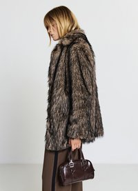 Manteau en fausse fourrure dans des tons marron foncé et beige, avec un col haut et une coupe ample, associé à un petit sac à main en cuir marron.