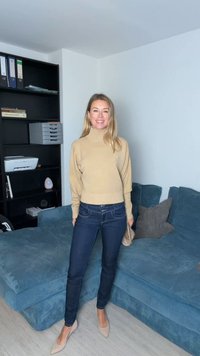 Camisola de gola alta bege, jeans azul escuro e saltos altos nude. O estilo inclui uma silhueta ajustada. O fundo apresenta um sofá azul.