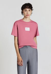 Persona che indossa una maglietta rosa con la scritta "ABBRACCIA TUTTO CIÒ CHE SEI" e pantaloni grigi larghi, in piedi contro uno sfondo chiaro e semplice.