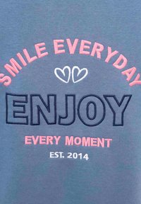 Grau gebleichtes Gewebe mit gesticktem Text: "LÄCHEL JEDEN TAG" in Rosa, "GENIEßEN" in Dunkelblau, "JEDEN MOMENT" in Rosa und "EST. 2014" in Weiß. Weiße Herzen zwischen den Worten.