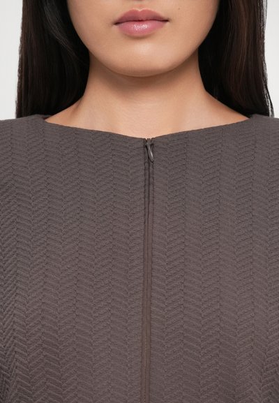 Brown textured garment with a chevron pattern, featuring a front zipper closure and a rounded neckline. The fabric appears ribbed.