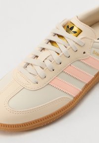 Baskets beige clair avec des bandes roses, dessus en cuir texturé et synthétique, design à lacets, semelle en caoutchouc naturel, détail du logo doré sur la languette.