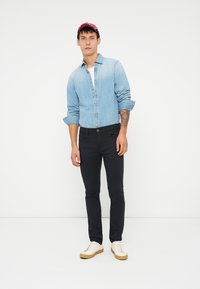 Ung mann iført rød caps, lyseblå denimskjorte, svarte bukser, hvite sneakers, med en tatovering på høyre underarm som står foran en hvit bakgrunn.