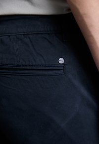 Gros plan d'une poche de pantalon bleu marine foncé avec un petit bouton métallique rond marqué « LONN » près de la ceinture.