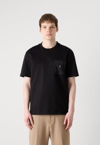 Czarna bawełniana koszulka z okrągłym dekoltem, krótkimi rękawami i teksturowaną kieszenią z logo. Nosi ją w połączeniu z beżowymi spodniami.