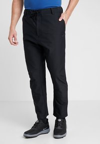 Černé sportovní kalhoty s páskem na stažení v pase, těsně střižené, s reflexním logem, v kombinaci s černými trekovými botami a modrým trikem.