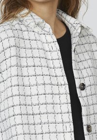 Hvid og sort ternet jakke i struktureret stof, med krave, to knapper og et fremtrædende rutenetmønster.