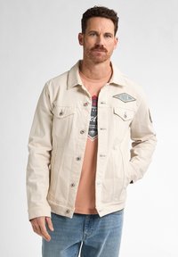 Chaqueta de mezclilla beige con frontal abotonado, bolsillos en el pecho y detalles de logotipo bordado. Lleva encima una camiseta rosa y unos jeans azules.