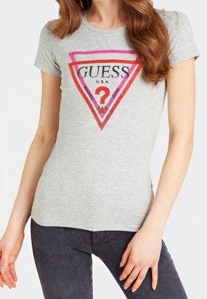 Femme portant un T-shirt gris ajusté avec un logo en triangle rouge et rose et le texte "GUESS U.S.A." sur le devant.