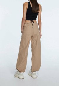 Pantalones cargo beige con dos bolsillos traseros, dobladillo ajustable con cordón, combinados con un top corto negro y zapatillas blancas. Acabado de tela mate.
