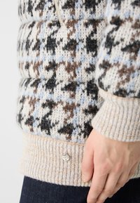Strickpullover mit einem gemischten Farbmuster aus Creme, Blau, Braun und Schwarz. Gerippter Saum und Bündchen mit einem kleinen metallischen Logodetail.