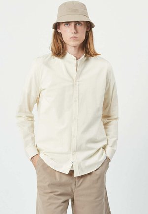 Camisa de manga larga beige con botones, hecha de una tela suave, que presenta un cuello mao y una parte delantera con botones. Llevada con pantalones cortos de color beige claro.
